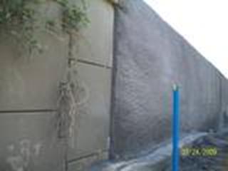 The photo shows a geosynthetic reinforced soil (GRS) wall built with concrete masonry unit (CMU) blocks. A layer of shotcrete has been sprayed over the face of the CMU blocks. The GRS wall is adjacent to a failed panel-faced mechanically stabilized earth (MSE) wall with vegetation growing through the joints of the panels. There is also blue inclinometer tubing extending in front of the MSE wall to monitor slope stability.