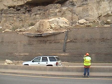 The photo shows a geosynthetic reinforced soil (GRS) wall that has been impacted by a very large rock from the top of a cliff overhang. The GRS wall is damaged at the top several courses but is still stable and standing. There is a man in a safety vest surveying the damage. He is standing on the other side of the barrier to the interstate. A white SUV is in front of the impacted wall.