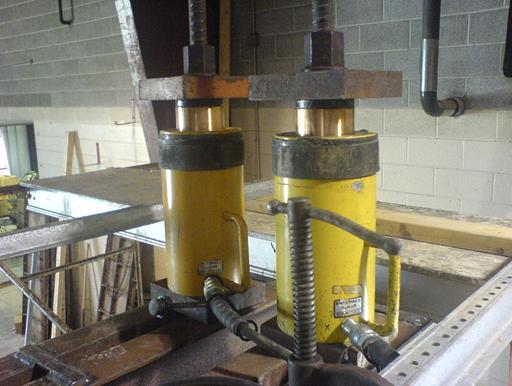 Photo showing two yellow hollow-core hydraulic jacks bolted to the top channel beams.