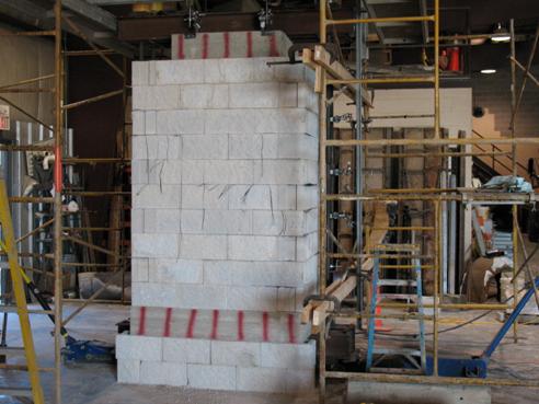 Photo showing the Defiance County experiment before loading. The geosynthetic reinforced soil (GRS) mass has a concrete pad at the top (not extending to the outside block facing) and a concrete base pad (extending to the outside block facing). Both concrete pads have vertical red lines spray-painted on them. Underneath the concrete base pad are concrete masonry unit (CMU) blocks. Scaffolding is shown around the right side of the experiment. Instrumentation is located on the top and right side of the GRS mass.