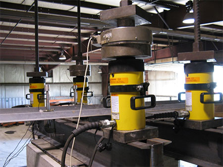 Figure 18. Photo. Hollow core hydraulic jacks for PT assembly. Four hydraulic jacks as viewed from a side angle comprise part of the performance test assembly for the Vegas mini-pier experiment. Hydraulic line connections can be seen on two jacks in the foreground and load cells are placed on top of the hydraulic jacks which are on top of the steel chanels, connected with a steel base plate.