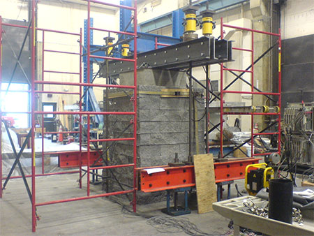 Figure 19. Photo. TF-1 PT set-up with reaction frame. Mini-pier test setup for test TF-1 from side angle in the Structures laboratory. Scaffolding is set up around the PT to serve as a stable platform for the lateral measurements. A concrete footing sits on top of the GRS composite with CMU facing elements. Steel channels rest on a steel plate on top of the concrete footing. Four hydraulic rams are located on top of the steel channels through a threaded bar attached to the strong floor.