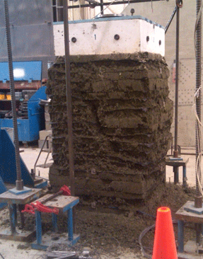 Figure 34. Photo. TF-11 at failure with Sv = 3-13/16 inches, Tf = 1,400 lb/ft, and 21A material. Side view of pier failure for test TF-11, with S subscript v equals 3-13/16 inches, t subscript f equals 1,400 lbs per ft, using 21A material. Layers are slumped and loose aggregate is strewn on the floor of the test facility. A steep shear failure plane is shown through the composite.