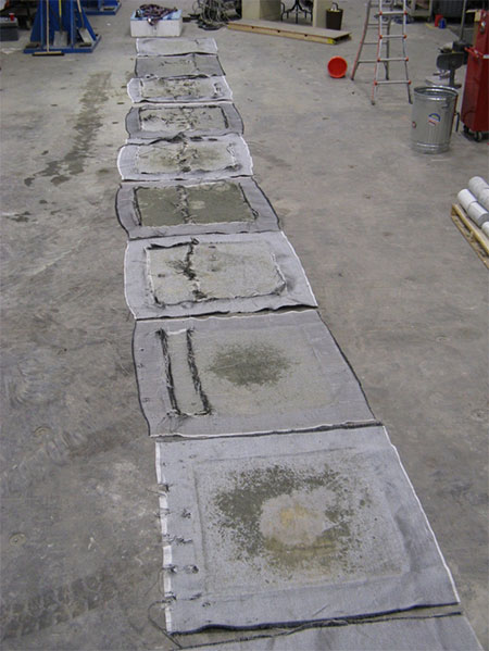 Figure 38. Photo. Rupture pattern for geotextiles in TF-6 (qult,emp = 43,828 psf); the lowest layer of reinforcement is the closet fabric in the picture. A line of geotextile squares used in test TF-6 is laid out in a line going away from the camera. In this test, q subscript ult, emp equals 43,828 psf. The lowest layer of reinforcement is the closet fabric in the picture.