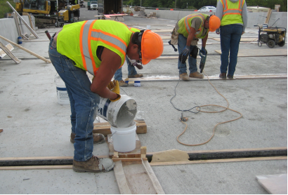 This photo shows the construction of field-cast ultra-high performance concrete (UHPC) deck-level connections. The UHPC is being poured into a bucket that is maintaining the pressure head on a connection space that has already been filled.