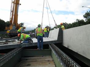 This photo shows the installation of a precast concrete deck panel with an integral concrete barrier. The panel is being placed over the exterior and first interior girders. It is adjacent to a parallel bridge span whose deck has already been replaced.