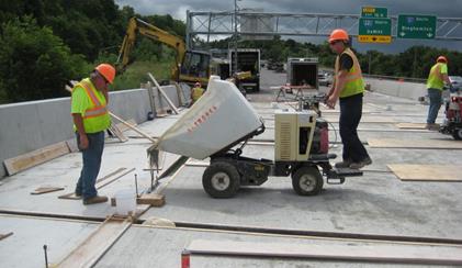 This photograph shows ultra-high performance concrete being poured from a motorized buggy into a deck-level connection on a bridge deck near Syracuse, NY. One workman operates the buggy while another monitors the operation.