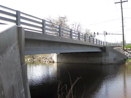 Figure 1. Photo. Route 31 bridge in Lyons, NY. This photograph shows the Route 31 bridge in Lyons, NY. It is the first bridge in the United States to use field-cast ultra-high performance concrete (UHPC) connections. The eight simple span deck-bulb-tee girders were joined through non-contact lap splice UHPC connections. An elevation view of the north side of the simple span bridge is shown, with the east abutment in the foreground and the west abutment in the background. The concrete girders span 86 ft (25 m) from abutment to abutment.