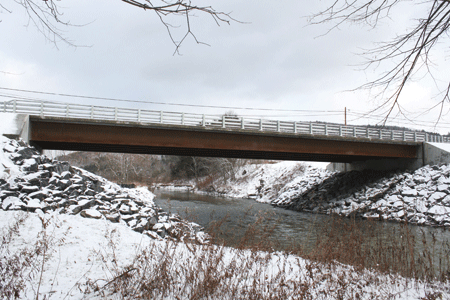 Figure 2. Photo. Route 23 bridge in Oneonta, NY. This photograph shows the Route 23 bridge in Oneonta, NY. This bridge is the second in the United States to use field-cast ultra-high performance concrete (UHPC) connections. It has precast concrete deck panels connected with field-cast UHPC connections. An elevation view of the south side of the simple span bridge is shown, with the west abutment on the left and the east abutment on the right. Steel girders span 128 ft (39 m) from abutment to abutment.  Riprap protects the structural concrete abutments on each bank of the creek.