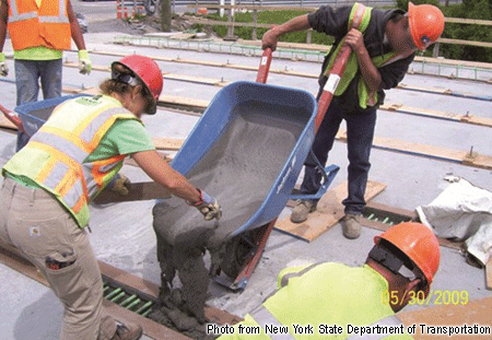 Figure 4. Photo. Longitudinal connections cast between deck-bulb-tee girders on the Route 31 bridge in Lyons, NY. This photograph shows workers casting fluid ultra-high performance concrete (UHPC) from a wheelbarrow into the void space between the top flanges of two deck-bulb-tee prestressed concrete girders. The rebar extends from the girders into the void. The UHPC is self-consolidating.