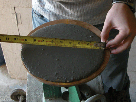 Figure 5. Photo. ASTM C1437 flow test on UHPC.(6) This photograph shows a rheology test being completed on ultra-high performance concrete (UHPC) immediately after mixing and before casting. There is a mini-slump cone filled with UHPC that is lifted, allowing UHPC to puddle. A person is seen measuring the width of the puddle with a measuring tape in inches.