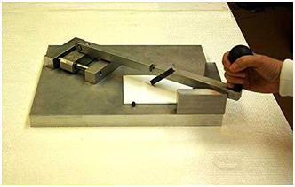 This photo shows a scribing tool made of stainless steel consisting of a 14.5- by 11- by 1-inch support base to rest the test panel while mechanically scribing it. The test panel is held in place by raised hinges. The surface of the base contains a guide rod fixture and a V-notch. A handle consisting of a scribing tip fixed at an angle slides back and forth on support guide rods to scribe the test panel. When the handle is not in motion, it rests in the V-notch.