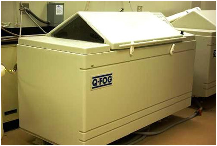This photo shows a salt-fog chamber, which consists of a unit to house the test panels while using cyclic corrosion of fog and dry-off processes. The unit is made of plastic and includes a custom controller to program the salt-fog and drying cycles. A solution reservoir, which feeds salt solution for the fog cycles, is next to the housing unit. Test panels can be loaded into the chamber from the top by opening a pyramid-shaped door. During the operation of the chamber, the door is secured using plastic hinges. The fog chamber is installed on the floor and cannot be moved.