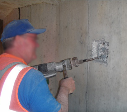 Figure 55. Photo. Concrete excavation to expose an internal tendon. This photo shows a worker with a power chisel excavating concrete on a vertical surface in order to access an internal tendon.