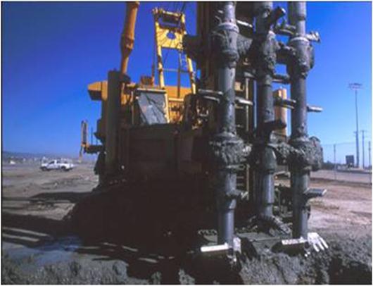 This photo shows wet rotary end (WRE) equipment for deep construction on land. Three axis mixing shafts are shown with a staggered arrangement of paddles at the ends of the shafts.