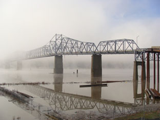 This picture shows the overall view of the bridge standing from the Indiana shore. Through the middle of the picture are six concrete piers protruding from the river. It is a foggy morning, and the furthest two piers and bridge sections over them are barely visible. The morning is still and the Ohio River is smooth as glass, showing a mirror-image reflection of the bridge in the bottom half of the figure. The extreme right middle of the picture shows the deck truss approach spans that transition to the through truss spans over the river through the middle of the photo.