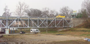 This photo focuses on just one of the approach span trusses, which takes up most of the width of the photo. The truss is simply supported and in a Pratt configuration. There is a white van parked beneath the bridge that demonstrates that the lower truss chord is approximately 15 ft off the ground. Atop the bridge is a yellow dump truck used as a controlled load; in the picture, it appears to be over the second node away from the Indiana shore.