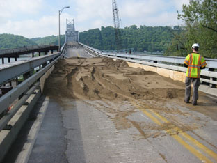 This photo is taken from the deck of the approach span deck truss on the Indiana side. In the background is the Kentucky shoreline, and the view is looking right down the middle of the bridge, so the through truss spans are also in the background. In the immediate foreground of the photo is sand, distributed evenly over the entire width of the truss and for about 52 ft along the truss. In the right foreground is a man in a yellow safety vest walking towards the sand.