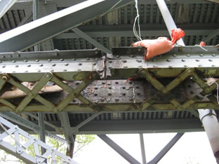 This photo was taken from the ground, looking up to the chord member after the first explosive event. The chord member is running left to right across the photo, and one of the two built-up C-shape elements of the member has been distinctly severed, leaving an approximate 1/2-inch gap where the shape charge was mounted. The background of the photo generally shows the bottom of the exodermic deck.