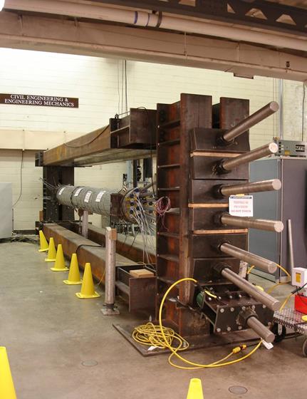 This photo shows a cable mockup representative of a full-scale section of a suspension bridge cable. The mockup cable is shown from the jacking end with all seven central strands tensioned. The threaded tensioning rods are seen with the hydraulic jacks and jacking beam in place on the first strand. The mockup cable is suspended carrying its own weight with only a small metal post placed at the center of the cable span. 