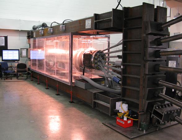 This photo shows a cable specimen in an environmental chamber. The mockup cable is again shown from the jacking end with the Plexiglas environmental chamber surrounding the specimen. The environmental chamber is seen mid-heat cycle with all three heat lamps functioning. The injection line for air-conditioning is shown on the nearside of the chamber. The heat lamp control instrumentation may be seen on one of the top beams of the surrounding mockup cable structure. 