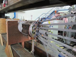 This photo shows the cable without the environmental chamber. The Plexiglas chamber has been removed from around the mockup cable. The aluminum wrapping has been removed from the surface of the cable. The image is taken from the jacking end with the tensioned strands and cable band in view. The cable is shown sitting in two of three wooden shoes built to hold the cable weight following the strand detensioning. 