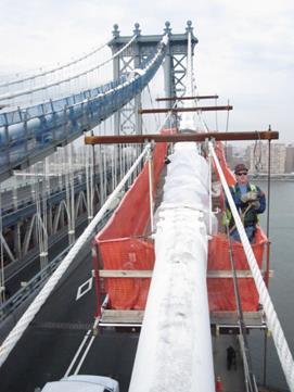 This photo shows a suspended platform between panel points 13 and 15 on the outer cable of the Manhattan Bridge. A worker stands on a suspended platform next to the north main cable section. The platform is built below the cable and is shown suspended from three cable bands and also attached to the guide wires. 