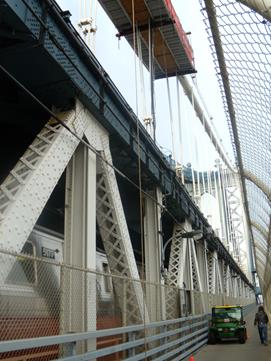 This photo shows a walkway where the data acquisition system (DAQ) was installed. The platform suspended below the north cable of the Manhattan Bridge is shown from the walkway below. The bridgeâ€™s truss structure and lower level is seen with a subway crossing on the external tracks. 