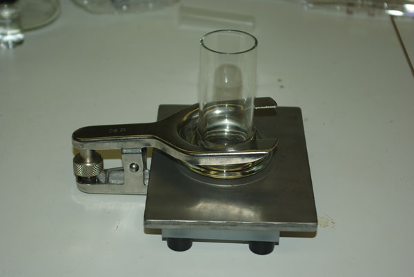 This figure shows a glass tube clamped onto a steel plate. The paint test cell is a glass tube with a rim on one end. A rubber O-ring is placed under the rim and the tube can be clamped on a flat surface. The rubber O-ring helps keep the tube watertight. The tube can hold 1.69 fluid ounces (50 milliliters) of water.