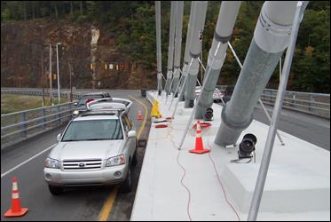 This photo was taken from on top of the median barrier on the side span of the bridge facing along the roadway. It shows the outer seven cables lined in a row and anchored into the median barrier in a way that one can observe the features of the cable, including the guide pipes, anti-vandalism pipes, neoprene boot, and HDPE connection sleeve, and the features of the damper, including the damper bracket and the steel damper support shaft. This photo was taken from on top of the median barrier on the side span of the bridge facing along the roadway. It shows the outer seven cables lined in a row and anchored into the median barrier in a way that one can observe the features of the cable, including the guide pipes, anti-vandalism pipes, neoprene boot, and HDPE connection sleeve, and the features of the damper, including the damper bracket and the steel damper support shaft.