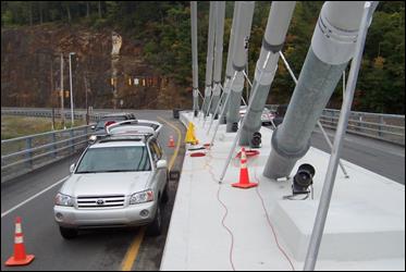 This photo was taken from on top of the median barrier on the side span of the bridge facing along the roadway. It shows the outer seven cables lined in a row and anchored into the median barrier in a way that one can observe the features of the cable, including the guide pipes, anti-vandalism pipes, neoprene boot, and HDPE connection sleeve, and the features of the damper, including the damper bracket and the steel damper support shaft.