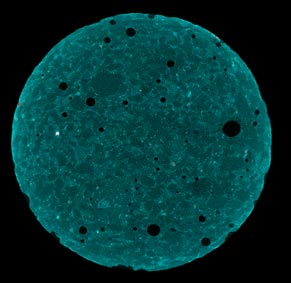 This photo shows an X-ray image of a cross-section of a circular sample before immersing it in 1 N NaOH solution. The pores are visible as a black spots on the surface of the sample. The unhydrated cement particles can be seen as white and grey speckles. The aggregate particles are gray-greenish in color, and the matrix is a light bluish-green in color. There are no signs of visible cracks.