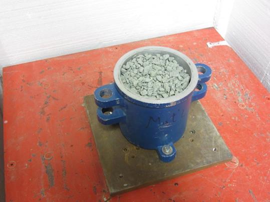 The photo shows a top view of a cylindrical 0.1 cubic ft mold filled with an open-graded aggregate, bolted to a vibratory table.
