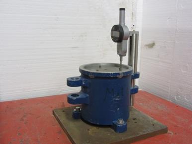 The photo shows a side view of a cylindrical 0.1 cubic ft mold bolted to a vibratory table with a digital linear variable differential transformer mounted on the top cover.