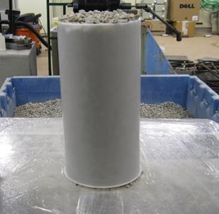 The photo shows a side view of a hollow 6 inch cylindrical plastic concrete mold filled with open-graded aggregate on top of a level plastic board. 