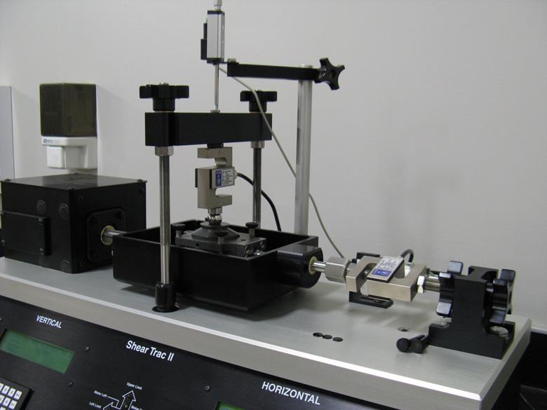 This photo shows a standard direct shear device. The device is automated with liquid crystal display panels and keypad buttons for vertical and horizontal control. The device has two load cells to measure the horizontal and vertical pressure. The device also has two linear variable differential transformers to monitor strain during the course of the experiment. The device has threaded rod clamps to secure the sample and a covered motor box with push rods to slowly shear the sample.