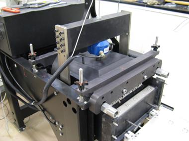 This photo shows the mechanical side of an automated large scale direct shear device. The soil sample bay setup for testing is shown along with the load cell to measure the vertical pressure on the top load plate directly over the soil. The top of the photo shows the tip of the linear variable differential transformer to monitor vertical strain during the course of the experiment. The photo shows many threaded connections to clamp the shear box together during the test.