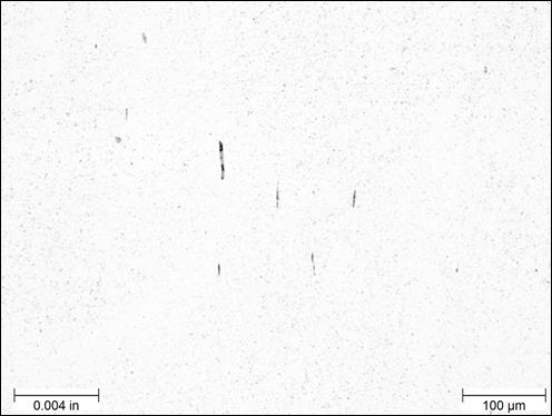 Figure 21. Photo. Unetched example of stringer inclusions near center of rod at 200x magnification. The micrograph was taken of an unetched sample near the center of the threaded rod viewed under bright field illumination at a magnification of 200x. A scale bar with a length of 0.004 inches is shown in the lower left corner of the photo, and a scale bar with a length of  100 micrometers is shown in the lower right corner. The micrograph is mostly a white background with small black spots interspersed throughout the photo. Five mostly black inclusions appear near the center of the photo. All five are elongated in the vertical direction, each having a width of approximately 0.0001 inches and lengths varying from approximately 0.0005 to 0.002 inches.