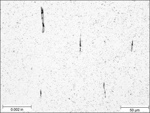 Figure 22. Photo. Unetched example of stringer inclusions near center of rod at 500x magnification. The micrograph was taken of an unetched sample near the center of the threaded rod, viewed under bright field illumination, at a magnification of 500x. A scale bar with a length of 0.002 inches is shown in the lower left corner of the photo, and a scale bar with a length of 50 micrometers is shown in the lower right corner. The micrograph is mostly a white background with small black spots interspersed throughout the photo. Five inclusions appear near the center of the photo. The inclusions are mostly black but have areas of gray as well. All five are elongated in the vertical direction, each having a width of approximately 0.0001 inches and lengths varying from approximately 0.0005 to 0.002 inches.