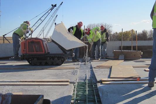 Figure 1. Photo. Casting of a grout-type material in the connections between PBEs. This photo shows the pour of a grout into the void space between prefabricated bridge elements (PBEs). The grout is pouring from a motorized buggy into an open strip between the reinforced concrete deck PBEs.