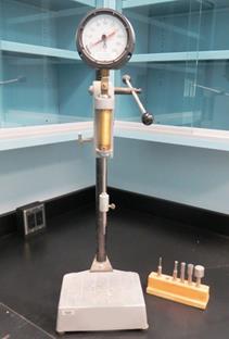 Figure 7. Photo. Loading apparatus and penetration needles. This photo shows the device used to measure the setting time of cementitious materials via the ASTM C403 test method. The device consists of a loading apparatus with a set of attachable standard flat-headed penetration needles of different sizes. The apparatus has a pressure gauge to measure the pressure force needed to force the needles to penetrate the fresh mixture. Based on the pressure read, the initial and final setting times of the material can be estimated.