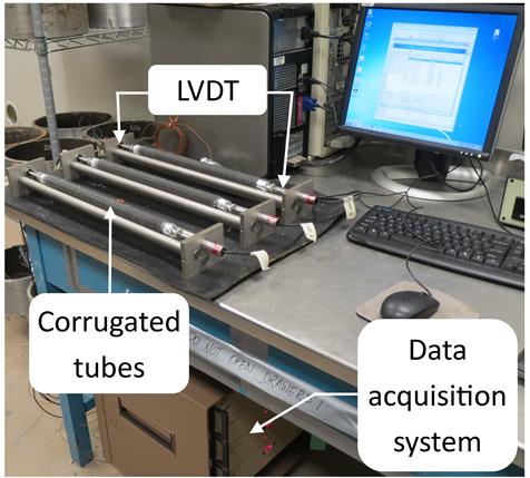 The photo shows the test setup to measure autogenous shrinkage on grout specimens according to ASTM C1698. The grout specimens consist of three corrugated tubes with a 1-inch (25.4-mm) diameter and about 17 inch (431.8-mm) length containing the fresh grout material. The tubes are sealed at both ends with plastic caps to avoid moisture loss to the environment. Each tube is placed on top of a rack that holds it in a horizontal position throughout the duration of the test. A linear variable differential transformer (LVDT) is located at each end of the rack so that the specimens’ length change over time can be measured. The LVDTs are connected to a data acquisition system as well as to a computer for the data collection.