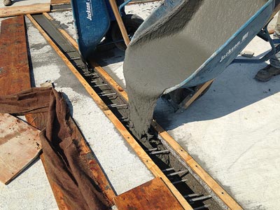 This photo shows the pour of a grout into the void space between prefabricated bridge elements (PBEs). The grout is pouring from a wheelbarrow into an open strip between the reinforced PBE concrete decks.