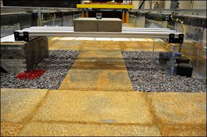 Figure 8. Photo. Riprap apron installation for testing. This photo shows the test set-up, viewed from upstream and looking downstream prior to the introduction of water. The colored rocks are at the upstream corner of the abutment on the left and the transparent abutment is located on the right.