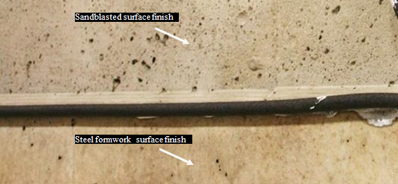 This photo shows the difference between two finishes on conventionally grouted connections. The surface of the connection in the top of the photo is sandblasted (SB). It appears to be rougher, with some of the air voids from the concrete exposed. The finish of the beam as it is removed from the framework is visible in the bottom half of the photo and appears smooth to the touch.