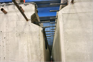 This photo shows beams with partial-depth connections being aligned to connect the ultra-high performance concrete (UHPC) shear key connection. One beam is resting in the testing configuration, and the second is suspended so it can be positioned adjacent to the stationary beam. Foam seals are visible on the bottom of the shear key connection and on the wooden spacers around the post-tensioning holes. Reinforcement protrudes from the connections of each of the beams.