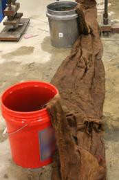 This photo shows wet burlap covering the freshly poured conventionally grouted connection. A continuous strip of burlap is seen running down the center of the connection. An additional roll of burlap is lying on top of the flat burlap. The rolled burlap is draped into 5-gal (18.925-L) buckets filled with water in order to keep the burlap wet.