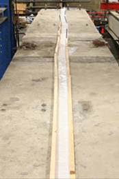 This photo shows a layer of white plastic sheeting covering the freshly poured ultra-high performance concrete (UHPC) connection. Plastic runs the entire length of the beam and is weighted down on either side of the connection with wood or reinforcing bars.