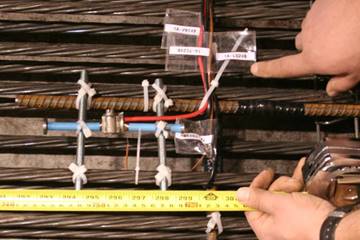 This photo shows a longitudinal embedded strain gauge and a thermocouple installed in the bottom flange of one of the beams prior to placing of concrete. The embedded strain gauge is aligned parallel to the prestressing strands. The thermocouple is directly next to this strain gauge as a brown wire. Red and blue wires are also shown as well as a ruler to provide context as to the length of the devices.