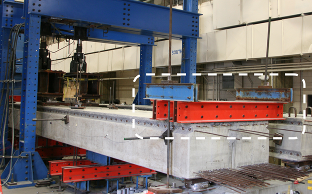 Figure 4. Photo. Clamping at the end of beams used in partially and fully stiffened cases. This photo highlights the clamping system that is used to reduce torsional rotation at the beam ends. The clamping system is highlighted in the photograph within a dashed rectangle. Two double channels are shown spanning transversely across the end of the beams. These steel channels serve to clamp down on the beam ends, attaching them to the strong floor through the use of threaded rods.